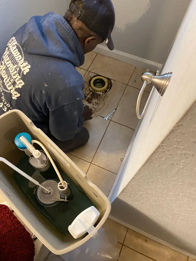 An Ellsworth technician in a company hoodie resetting a toilet flange, tank lid in the foreground